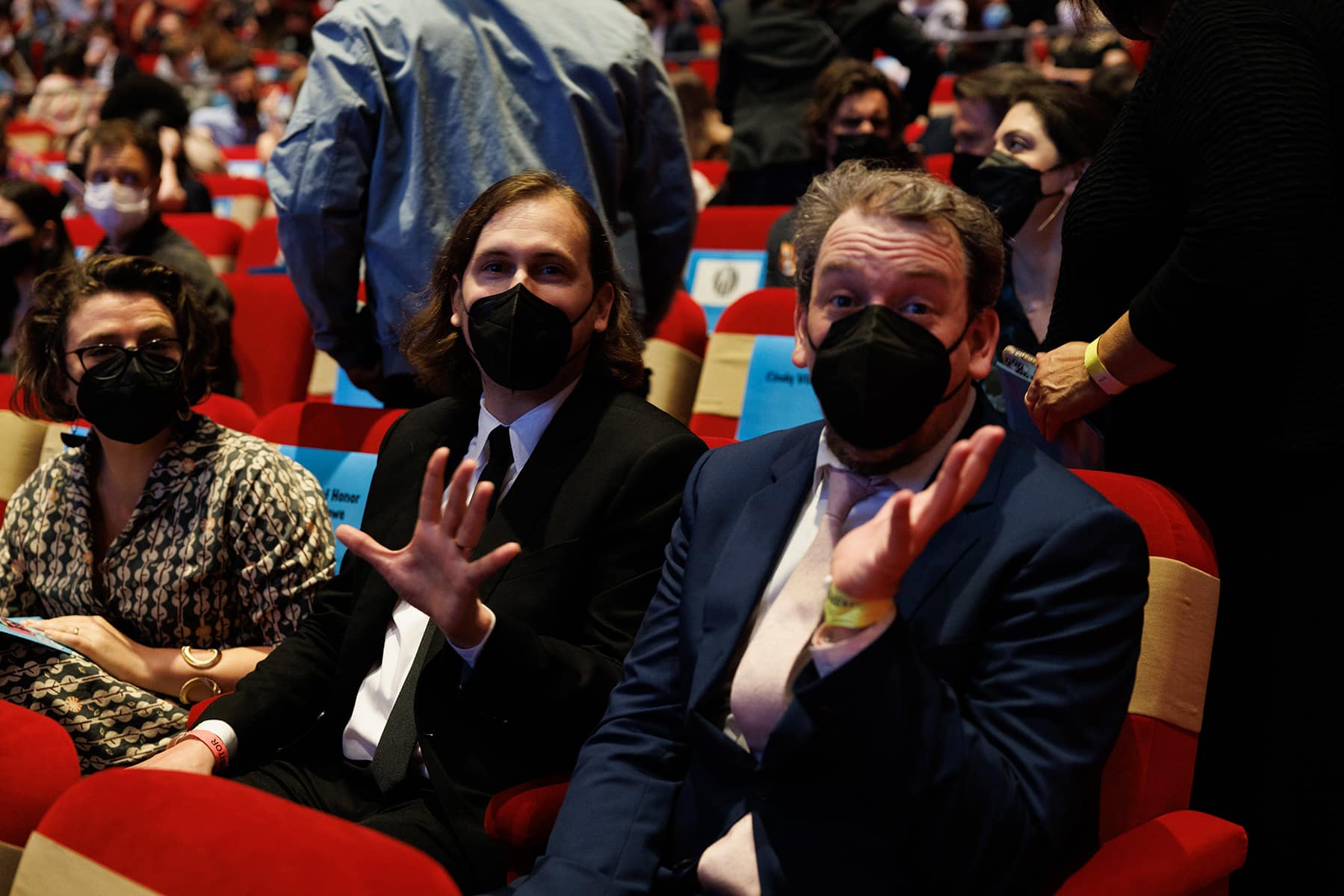 The two, seated and wearing masks, wave at the camera.