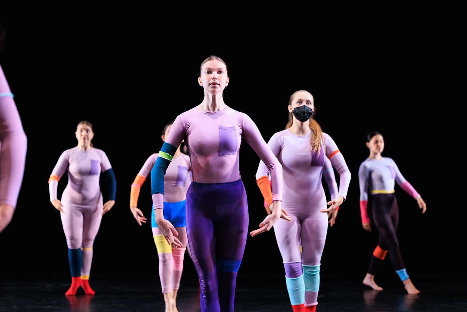 Array of dancers, wearing patchwork leotards, holding the same upright pose.
