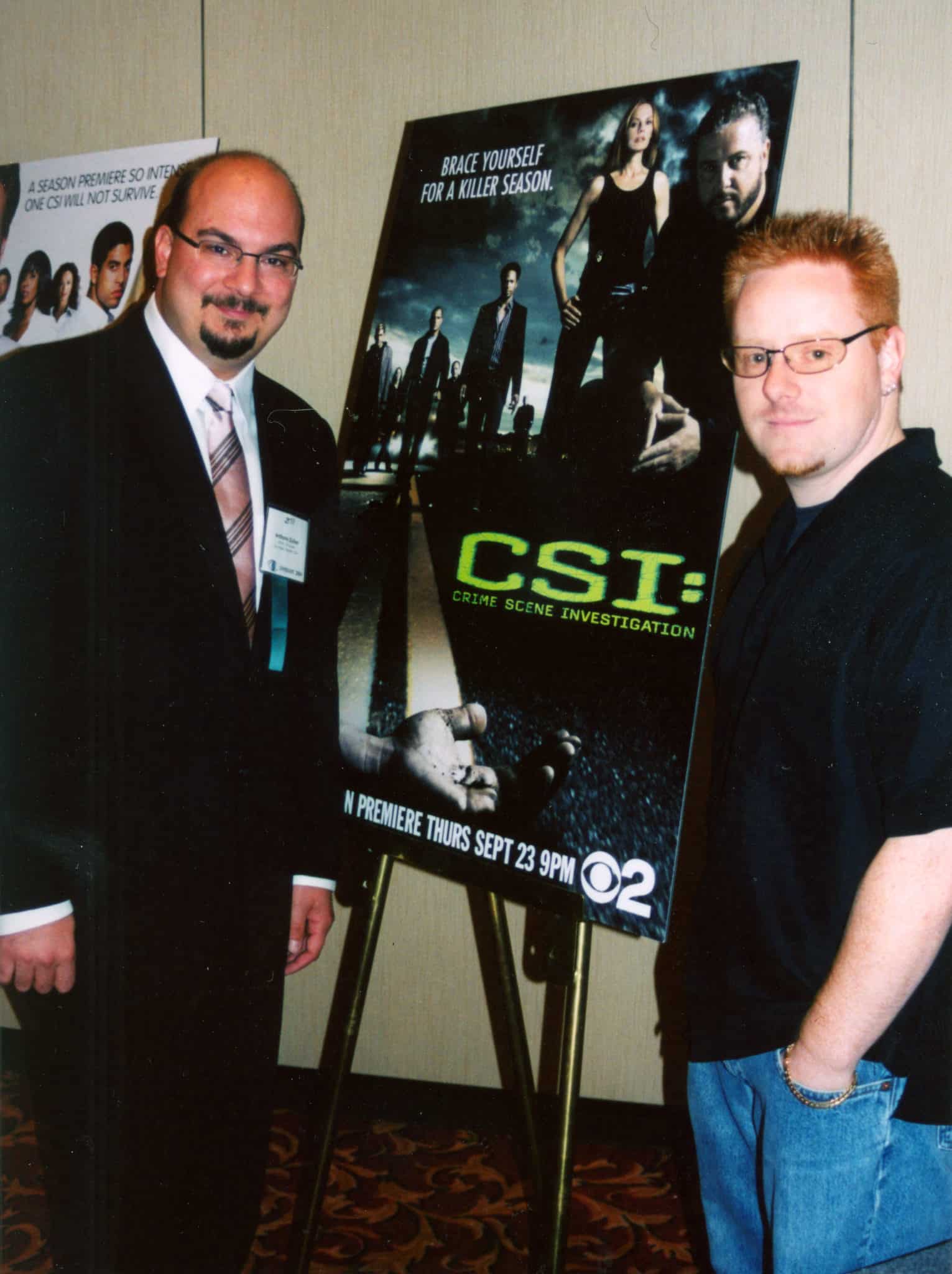 two men stand aside a poster for CSI tv show