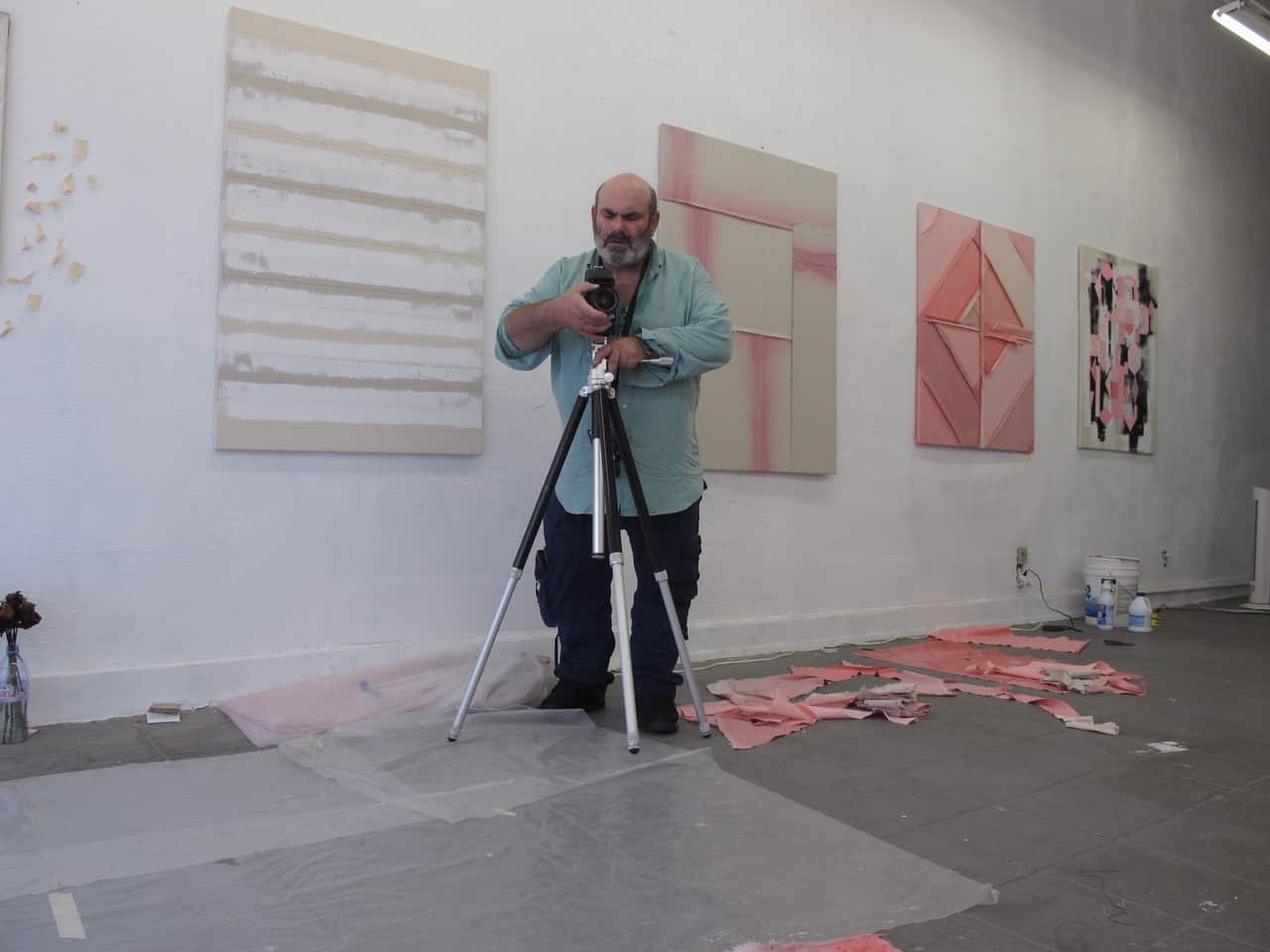 The artist setting a camera on a tripod. Behind him is a studio wall with contemporary artworks hanging.