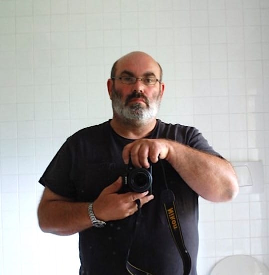 The artist, wearing a black T-shirt and glasses, holding a Nikon camera. Behind him is a white tiled wall.