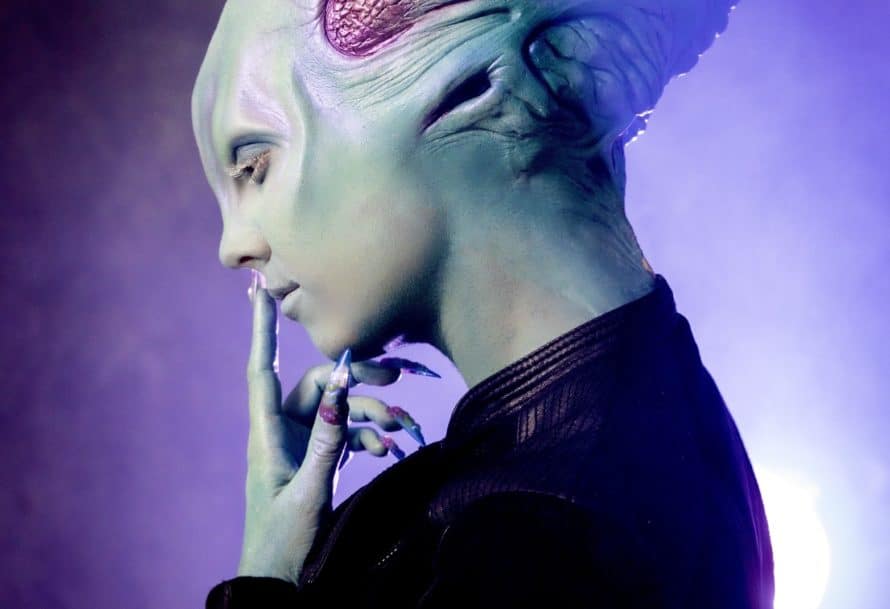 Side profile of person in alien mask and prosthetics bringing fingers to lips
