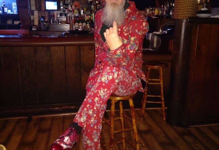 Man in a hat, sunglasses, and vibrant red floral patterned suit sits cross-legged on wooden bar stool