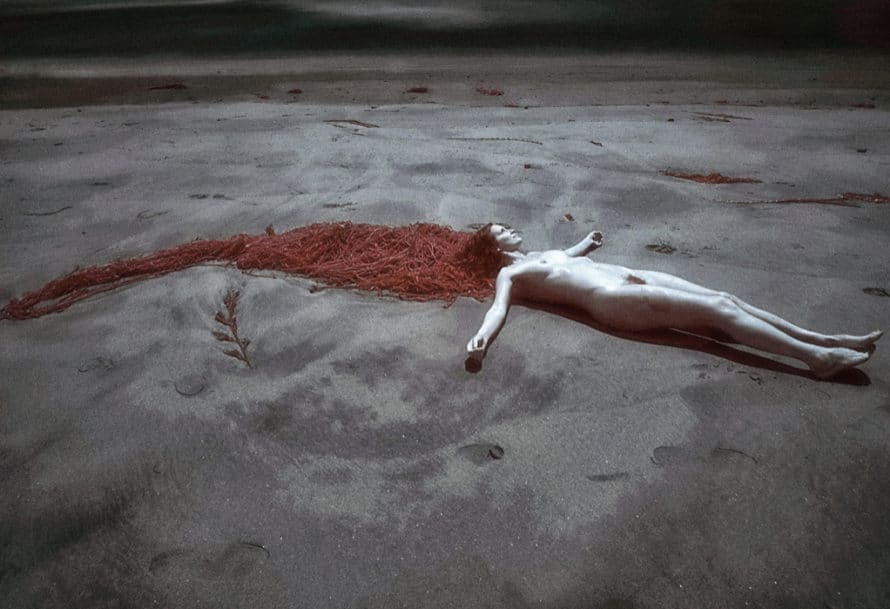 Nude woman lays head against pile of red cords on gray sand
