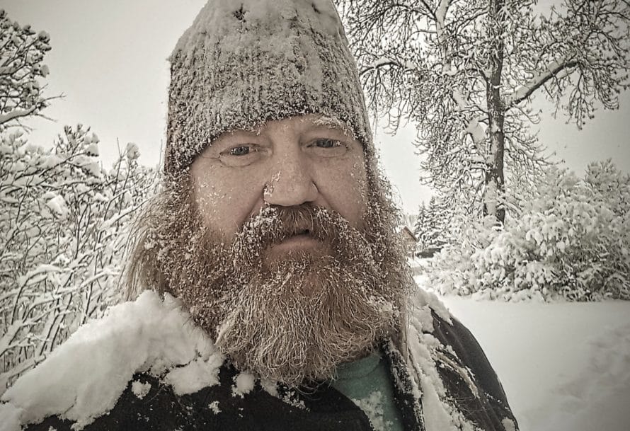 Bearded man in a hat dusted in snow standing against snowy white trees and sky