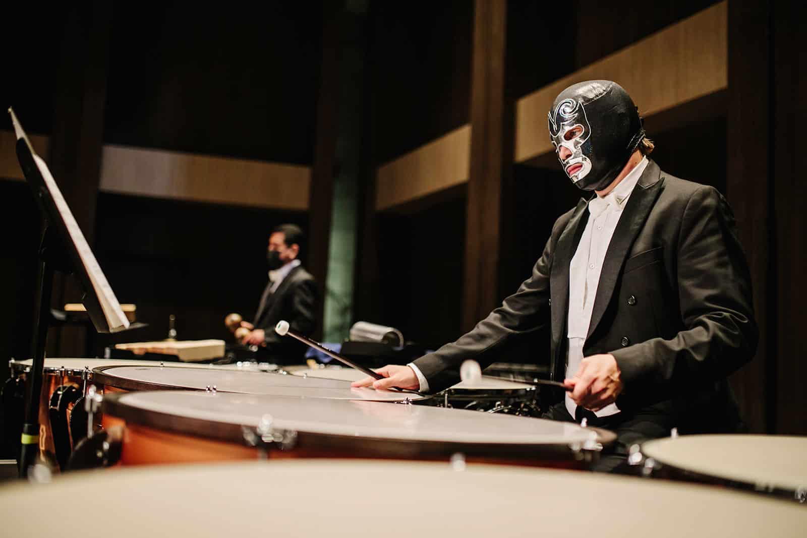 Person in black and silver luchador mask playing timpani drums