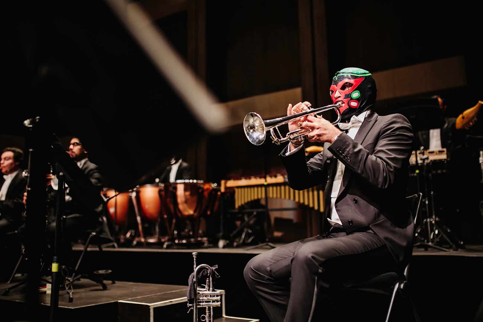 Seated trumpet player wearing black, green, and red luchador mask with orchestra