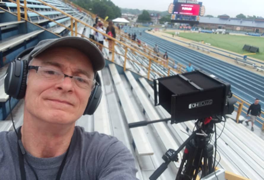 David L. Emerson on bleachers of sports arena, wearing headphones and standing next to camera equipment