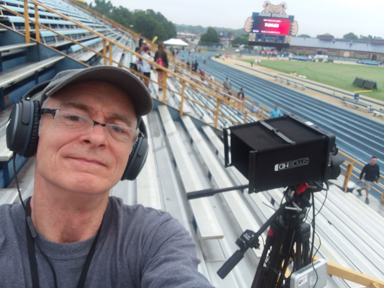 David L. Emerson on bleachers of sports arena, wearing headphones and standing next to camera equipment