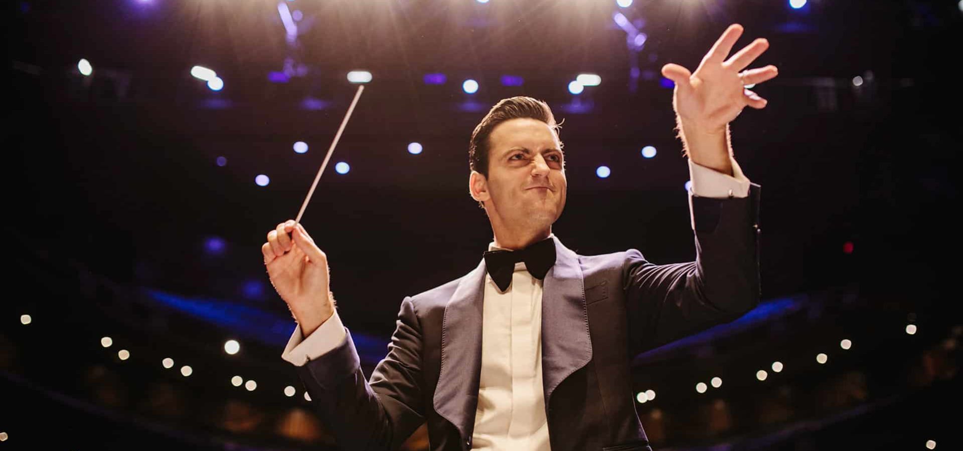 Conductor gestures at orchestra with left hand while holding baton in the right