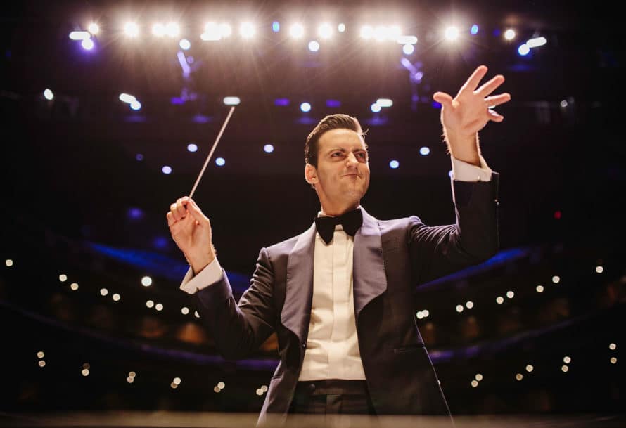Conductor gestures at orchestra with left hand while holding baton in the right
