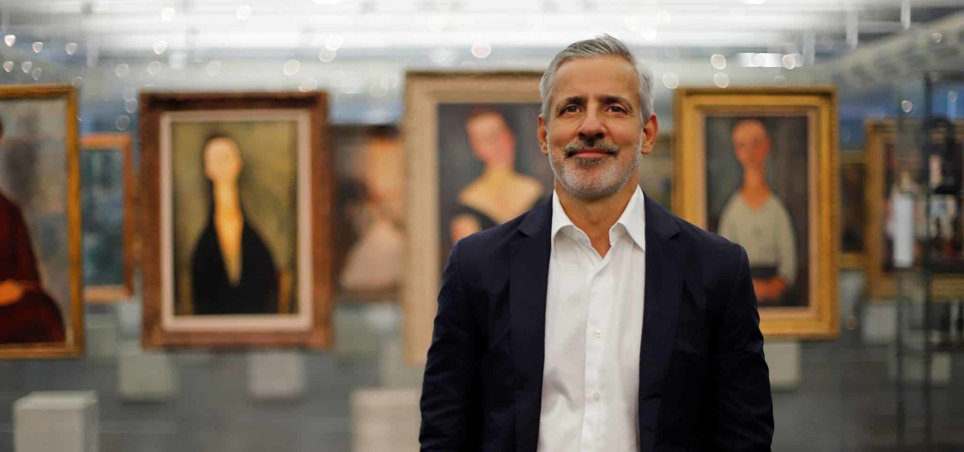 Adriano Pedrosa stands smiling in gallery setting featuring hanging paintings