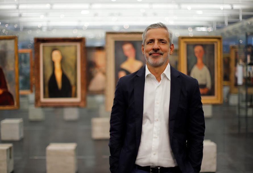Adriano Pedrosa stands smiling in gallery setting featuring hanging paintings