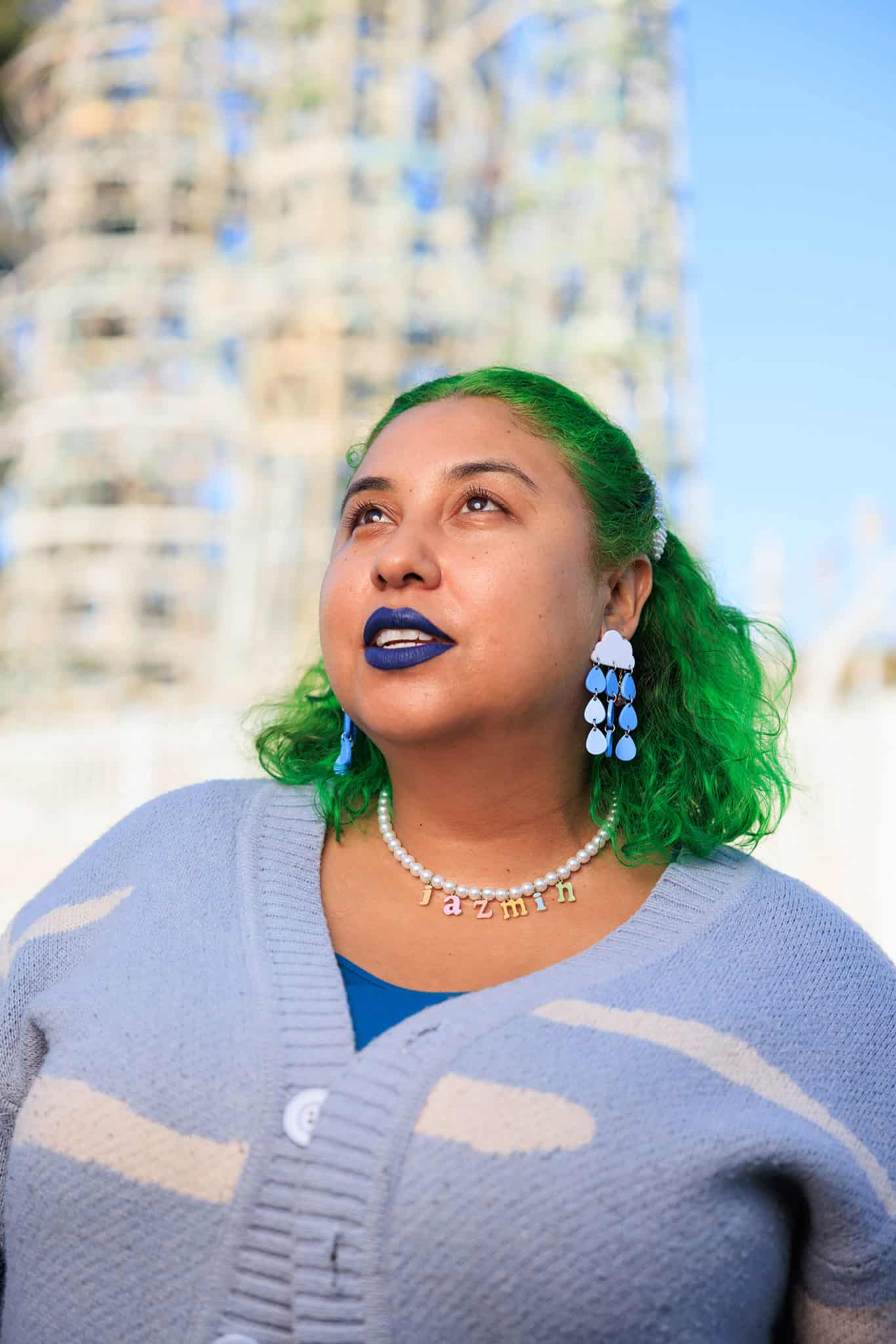 Jazmín, with bright green hair and raincloud earrings, looks skyward standing against Watt Towers