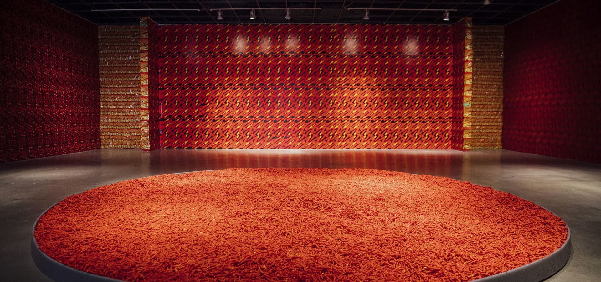 Gallery space with pool of Hot Cheetos on the floor and walls papered with hot cheeto bags