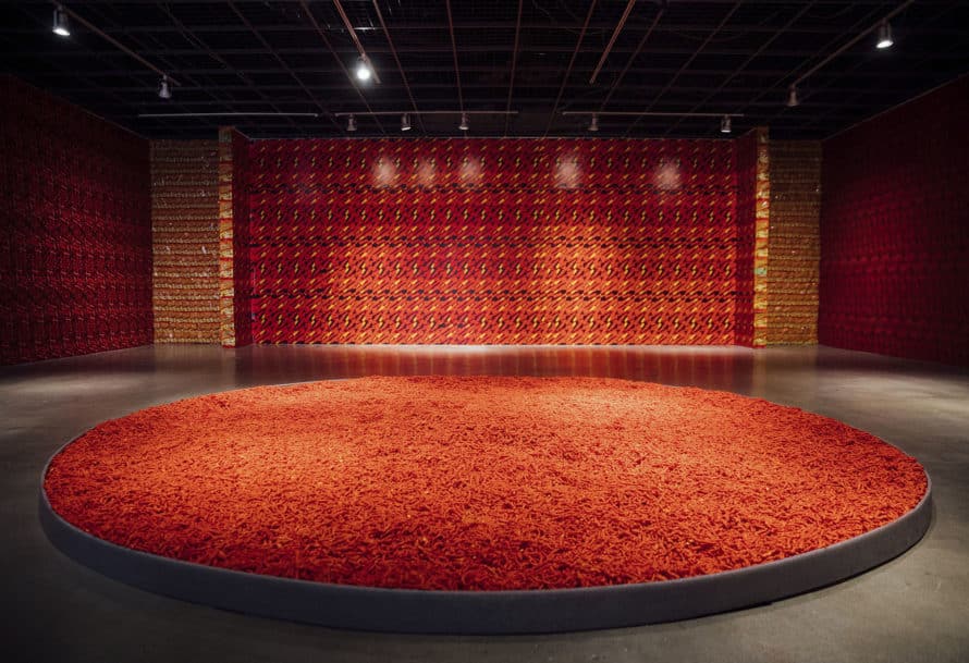 Gallery space with pool of Hot Cheetos on the floor and walls papered with hot cheeto bags