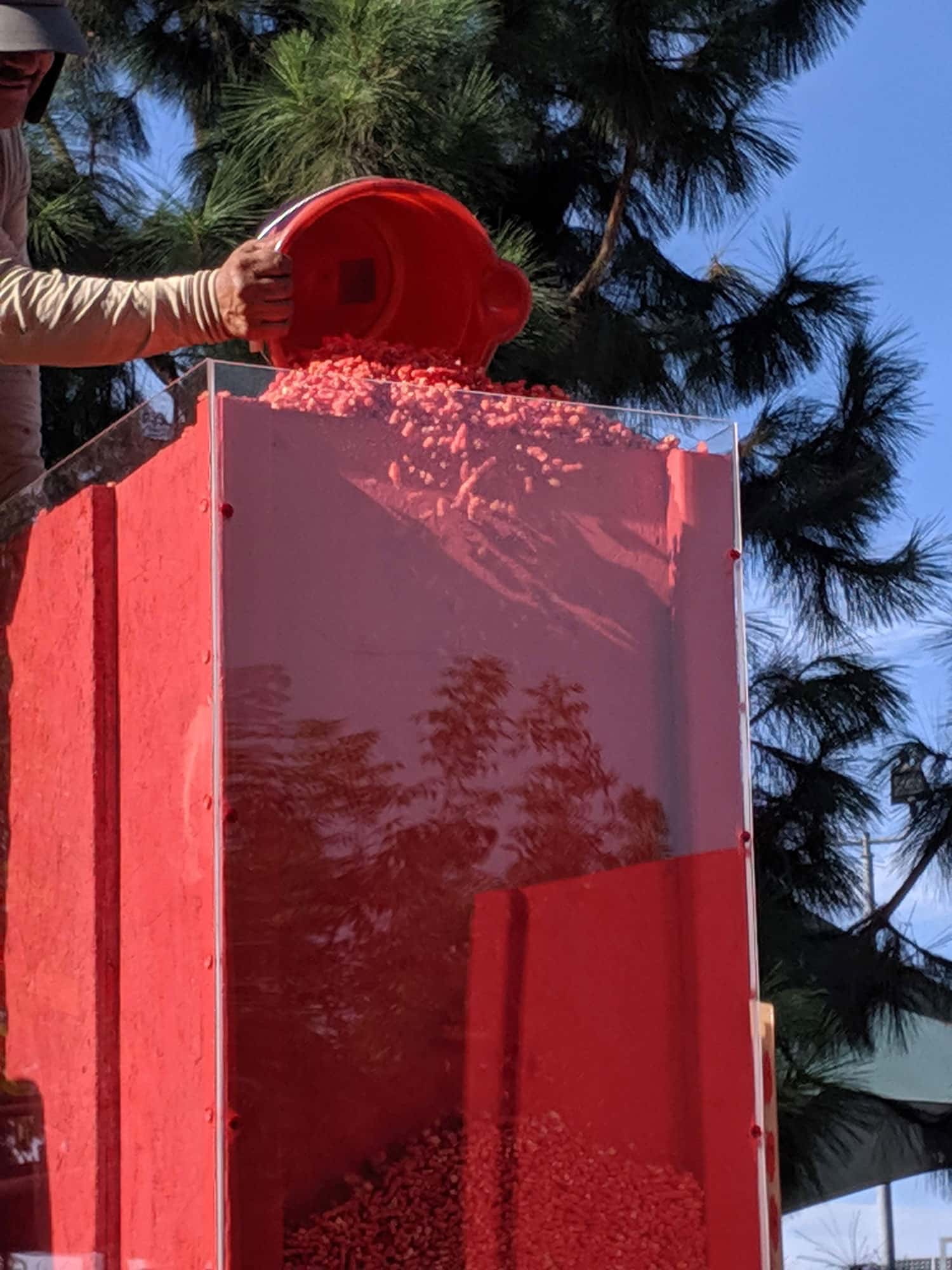 Person's gloved hand pours bucket of Hot Cheetos into large glass structure
