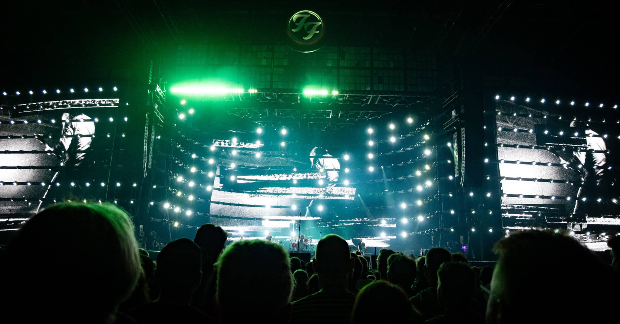 A concert scene featuring an audience in front of a brightly lit stage, showcasing a large screen with black and white visuals and vibrant green lights.