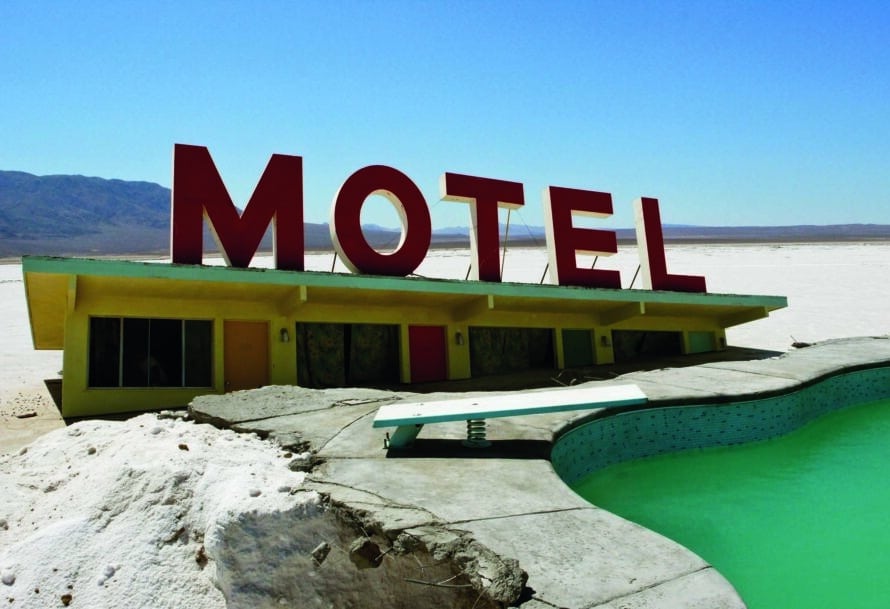 A vintage yellow motel with a large red “MOTEL” sign, an empty turquoise pool, and a cracked concrete patio in a barren landscape.