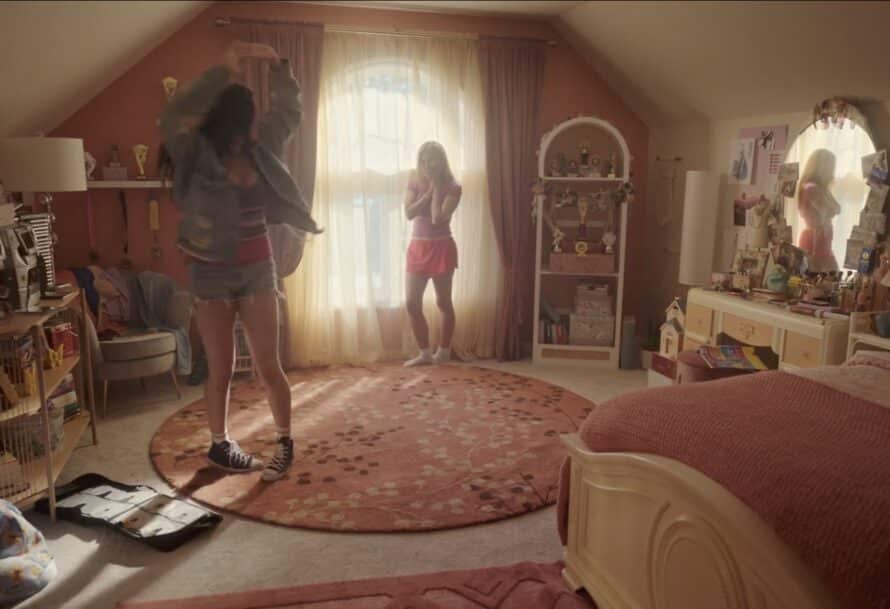Two young women in a bright bedroom; one spins in a denim jacket, while the other looks surprised. The room is decorated in warm colors, featuring a round orange rug and shelves with trophies.